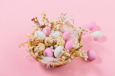 Easter candy chocolate eggs and almond sweets lying in a birds nest decorated with flowers and feathers on pink background. Happy Easter concept.