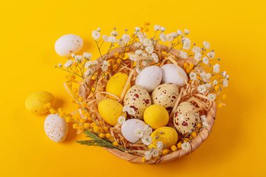 Easter candy chocolate eggs and almond sweets lying in a birds nest decorated with flowers and feathers on a yellow background. Happy Easter concept.