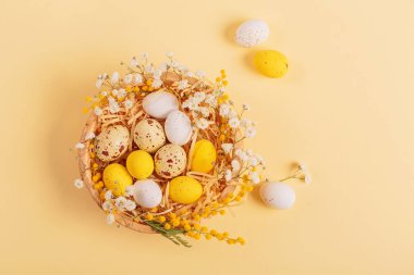 Easter candy chocolate eggs and almond sweets lying in a birds nest decorated with flowers and feathers on a yellow background. Top view. Happy Easter concept.