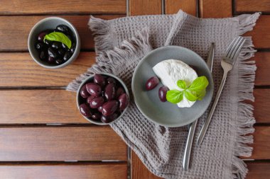 Gri tabakta dilimlenmiş Feta peyniri, siyah zeytin ve fesleğen yapraklarıyla. Terasta lezzetli ve sağlıklı bir kahvaltı.