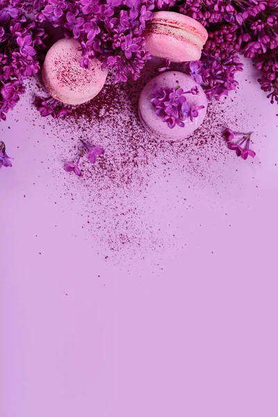 Pastel colored sweet french macaroons with lilac flowers and splash of dry blueberry powder on pink background. Beautiful composition for bakery and pastry shop. Top view with copy space