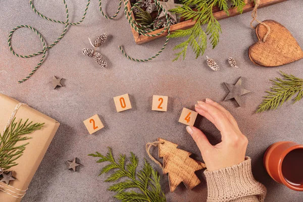 Winter holiday mood, Merry Christmas, Happy New Year background. Preparation for holidays. Handmade gift box, wooden box with Christmas decor and womans hand adding up numbers 2024. Flat lay, top view