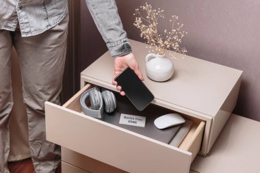 Aygıtsız Bölge etiketli ayrı masa çekmecesinde akıllı telefonu tutan el kamerasını kapat. Kadın telefonunu farklı aletlerle çekmecenin içine koyuyor. Dijital detoks ve teknoloji bağımlılığı konsepti