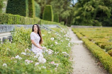 Portrait of young attractive woman sitting among white roses in picturesque old park of small town near an ancient French castle