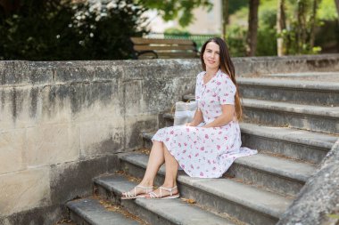 Attractive young woman with long brunette hair sitting and posing with big shopping bag on ancient stone stairs outdoor in old french city