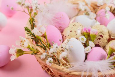 Easter candy chocolate eggs and almond sweets lying in a birds nest decorated with flowers and feathers on pink background. Happy Easter concept.