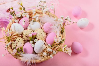 Easter candy chocolate eggs and almond sweets lying in a birds nest decorated with flowers and feathers on pink background. Happy Easter concept.
