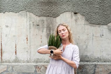 Attractive blonde woman with eyes closed in pink dress holding plant in pot and craft paper in her hands