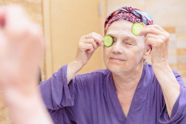Olgun bir kadın yüzüne bakıyor. Banyoda aynanın karşısında duran ve yüzüne krem ya da maske süren ve gözlerinde salatalık dilimleri olan son sınıf öğrencisi bir kadın.