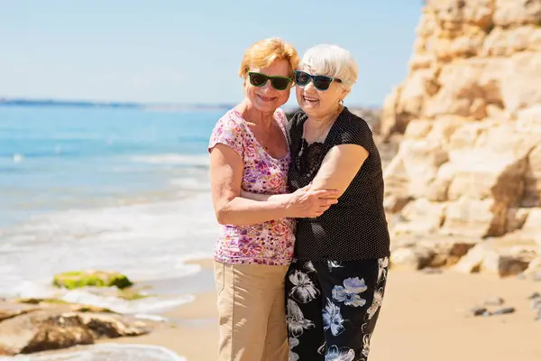 Two senior women are metting on a walk along a rocky beach, talking, hugging and laughing, having a pleasant time together.