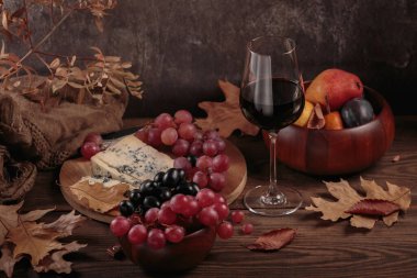 Sonbahar sıcacık hayatı, bir bardak kırmızı şarap, peynir tabağı mevsimlik meyveler ve koyu ahşap arka planda kırsal tarzda kuru yapraklar. Şarap tatma, kazak hava durumu konsepti.