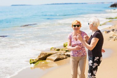İki yaşlı kadın kayalık sahilde yürüyor, konuşuyor ve gülüyor, birlikte hoş vakit geçiriyorlar.