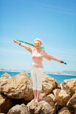 Mutlu olgun kadın gülümsüyor ve kumsalda eğleniyor. Sahilde mavi bir eşarpla yürüyen yaşlı bir kadının yaz portresi.