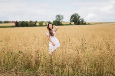 A beautiful brunette woman in a white dress runs along a field of golden chickpeas ear. Stylish girl in the field in windy weather