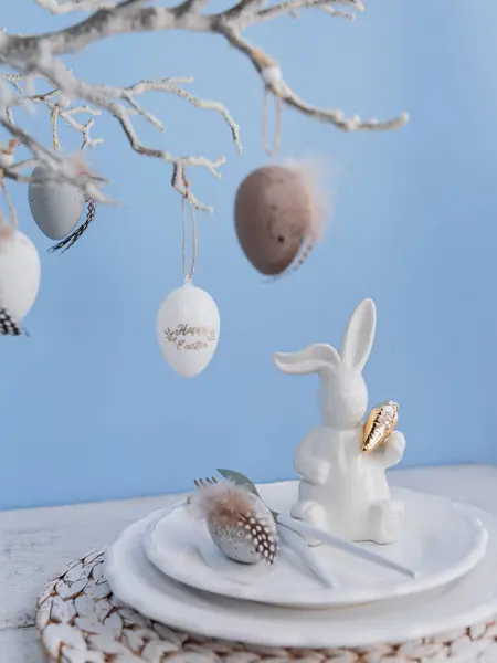 Table place setting with white bunny figure and eggs and festive easter decor on pastel blue background. Happy easter greeting or invitation card