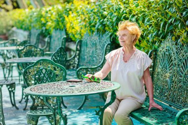 Mature attractive woman traveler sitting alone on the terrace of coffee shop in beautiful park in Bulgaria. Active life of the elderly in retirement, active seniors