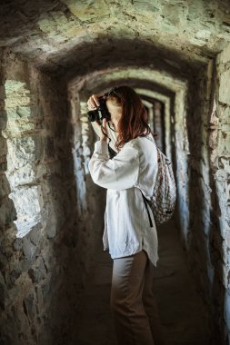 İlgilenen kadın turist, antik şatonun taş koridorunda dikilirken fotoğraf makinesinde fotoğraf çekiyor.