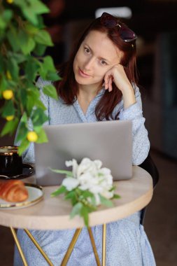 Masada oturan, kahve ve kruvasan içerken dizüstü bilgisayar kullanan mavi elbiseli çekici bir kadın.