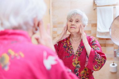 Mature woman taking care of her face. Senior woman standing at the mirror in the bathroom and applying cream to her face or doing self-massage