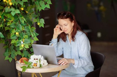 Masada oturan, kahve ve kruvasan içerken dizüstü bilgisayar kullanan mavi elbiseli çekici bir kadın.