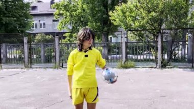Sarı futbol kıyafetli genç bir çocuk şehir sahasında top kontrolü yapıyor, kendini adamışlık ve atletizm gösteriyor..