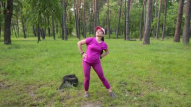 Pembe spor kıyafetli olgun bir kadın yeşil bir parkta egzersiz yapıyor, açık hava eğitiminden zevk alıyor. Yüksek kaliteli FullHD görüntüler