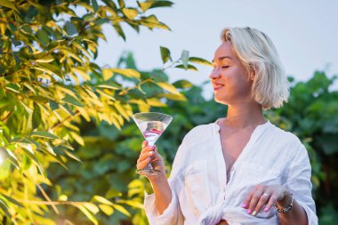 Attractive young blonde woman drinking dry martini in the backyard in the evening. Cocktail outdoors party time