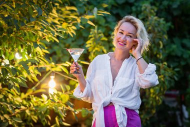 Attractive young blonde woman drinking dry martini in the backyard in the evening. Cocktail outdoors party time