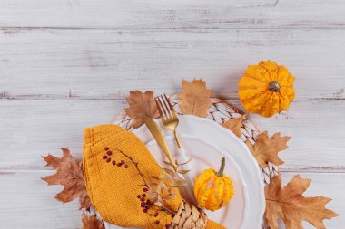Beyaz ahşap arka planda balkabağı süslemeleri olan sonbahar masası. Boş tabak, çatal bıçak takımı, sarı keten peçete ve kuru yapraklar. Sonbahar tatili, Cadılar Bayramı, Şükran Günü konsepti. Üst görünüm