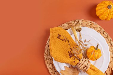 Turuncu arka planda balkabağı süslemeleri olan sonbahar masası. Boş tabak, çatal bıçak takımı, sarı keten peçete ve kuru yapraklar. Sonbahar tatili, Cadılar Bayramı, Şükran Günü konsepti. Üst manzara, düz uzanma