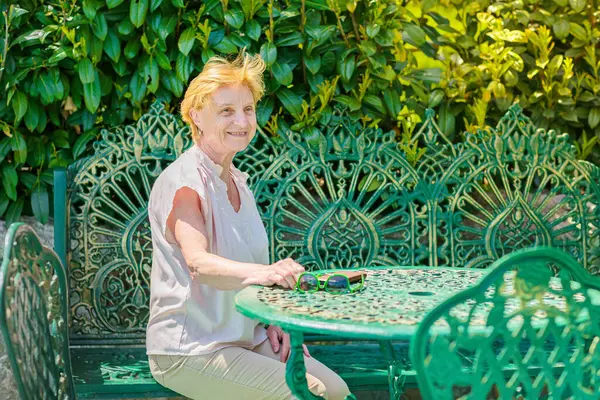 Mature attractive woman traveler sitting alone on the terrace of coffee shop in beautiful park in Bulgaria. Active life of the elderly in retirement, active seniors