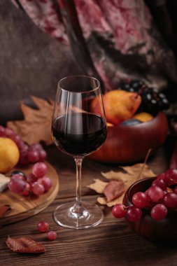 Koyu ahşap arka planda meyvelerle servis edilen bir bardak zengin kırmızı şarap. Sonbahar, şarap, üzüm ve kuru yapraklarla dolu kırsal bir hayat. Süveter hava konsepti