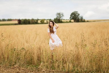 Young woman in white midi dress walking across the field with ripe golden ears of chickpeas. Beauty Romantic woman Outdoors