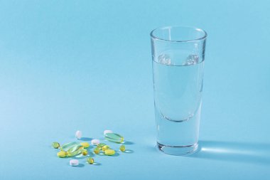 Diyet, beslenme, sağlıklı yeme konsepti. Çeşitli ilaç hapları, haplar, kapsüller ve mavi arka planda akan bir bardak su.