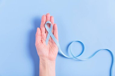 Mavi bir kanser kurdelesini tutan bekar bir kadının eli açık mavi bir arka planda dinleniyor. Kolorektal kanser hastaları ve ilgili kampanyaların farkındalığını ve bakımını simgeliyor.