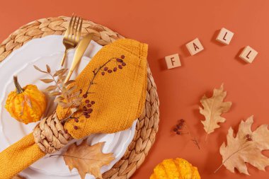 Balkabağı süslemeleri ve turuncu arkaplandaki yazıtlarla sonbahar masası ayarı. Boş tabak, sarı peçete ve kuru yapraklar. Sonbahar tatili, Cadılar Bayramı, Şükran Günü konsepti. Üst görünüm