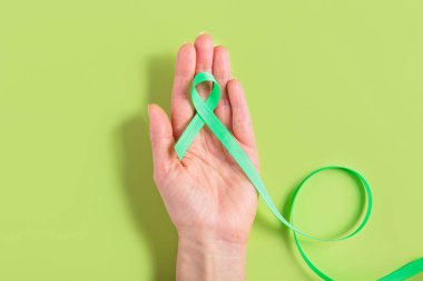 Bir kadının elinde tutulan yeşil farkındalık şeridi eşleşen yeşil arka plana karşı karaciğer kanseri farkındalığını sembolize ediyor. Sağlık kampanyaları ve savunma girişimleri için ideal.