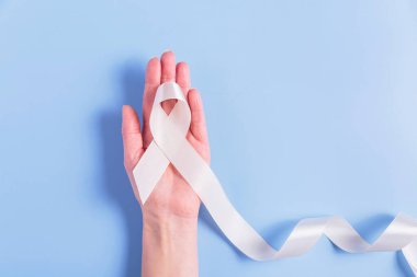 Beyaz bir kurdele, bir kadının elinde mavi bir arka plana karşı nazikçe tutularak kemik kanseri için destek ve farkındalığı sembolize ediyor. Tıbbi promosyonlar ve hasta desteği için ideal..