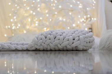 Yumuşak, tıknaz beyaz örgü, yansıtıcı bir yüzey üzerinde düz uzanır. Arka planda sıcak, parıldayan bokeh ışıkları, huzurlu ve rahat bir tatil atmosferi yaratır..