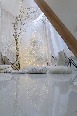 Zarif örgü battaniyeler, kabarık yastıklar ve minimalist mobilyalar ile çevrili, peri ışıklarıyla süslenmiş beyaz bir Noel ağacının yer aldığı şık bir iç mekan..