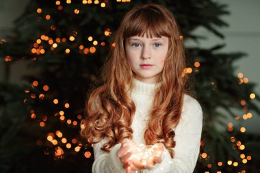 Evin rahat oturma odasında Noel ağacının yanında oturan ve elinde çelenk tutan mutlu genç kız. İyi kış tatilleri.