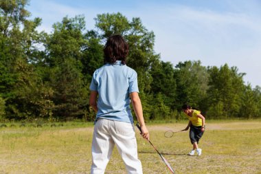 Kadın ve genç çocuk güneşli bir çimenlikte badminton oynuyorlar.