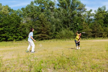 Olgun kadın ve genç çocuk güneşli bir günde çimenli bir tarlada badminton oynuyorlar.
