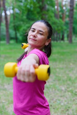 Yeşil bir parkta spor yaparken iki kolunu da dambılla kaldıran olgun bir kadın. Güç ve zindeliğe odaklanıyor..