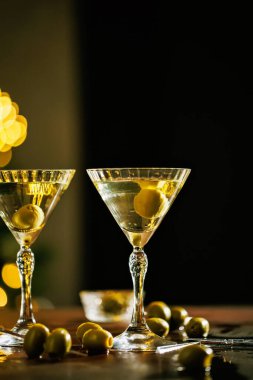 Temiz bir kokteyl ve yeşil zeytinle dolu sofistike bir martini bardağı, altın bokeh ışıklarının göz kamaştırıcı arka planına kurulmuş, neşeli bir havayı çağrıştırıyor..
