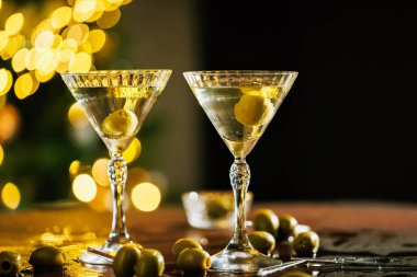 Yeşil zeytinli bir çift martini bardağı, karanlık bir masanın üzerine yerleştirilmiş, sıcak altın tatil ışıklarıyla dolu, rahat ve lüks bir atmosfer yaratıyor..