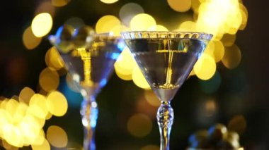 Bir kadın eli zarif bir şekilde martini bardağına zeytin garnitürü koyar, ılık, ışıldayan bir bokeh arka planına kurulur..