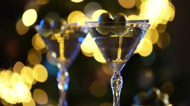 İki zeytinli martini bardağı, arka planda sıcak altın bokeh olan ahşap bir masanın üzerine yerleştirilmiş..