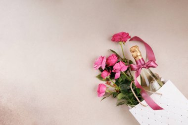Pembe kurdeleli bir şişe şampanya ve bej arka planda beyaz bir hediye çantasında taze pembe güller..