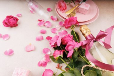Taze pembe güller, saten kurdeleli şampanya, narin taç yaprakları ve altın sofra takımı içeren romantik bir masa..